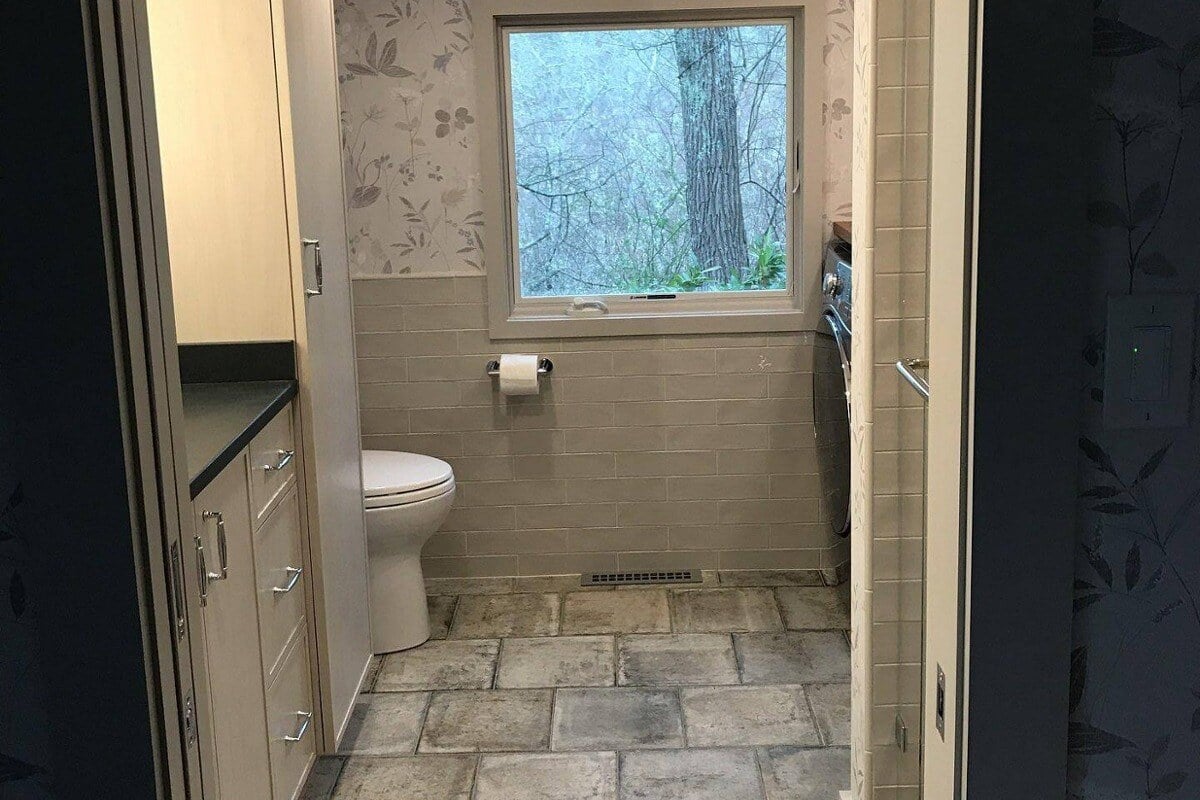 Bright bathroom with stone tile flooring and natural light from a large window in a Cape Cod home remodeled by @designREMODEL-1